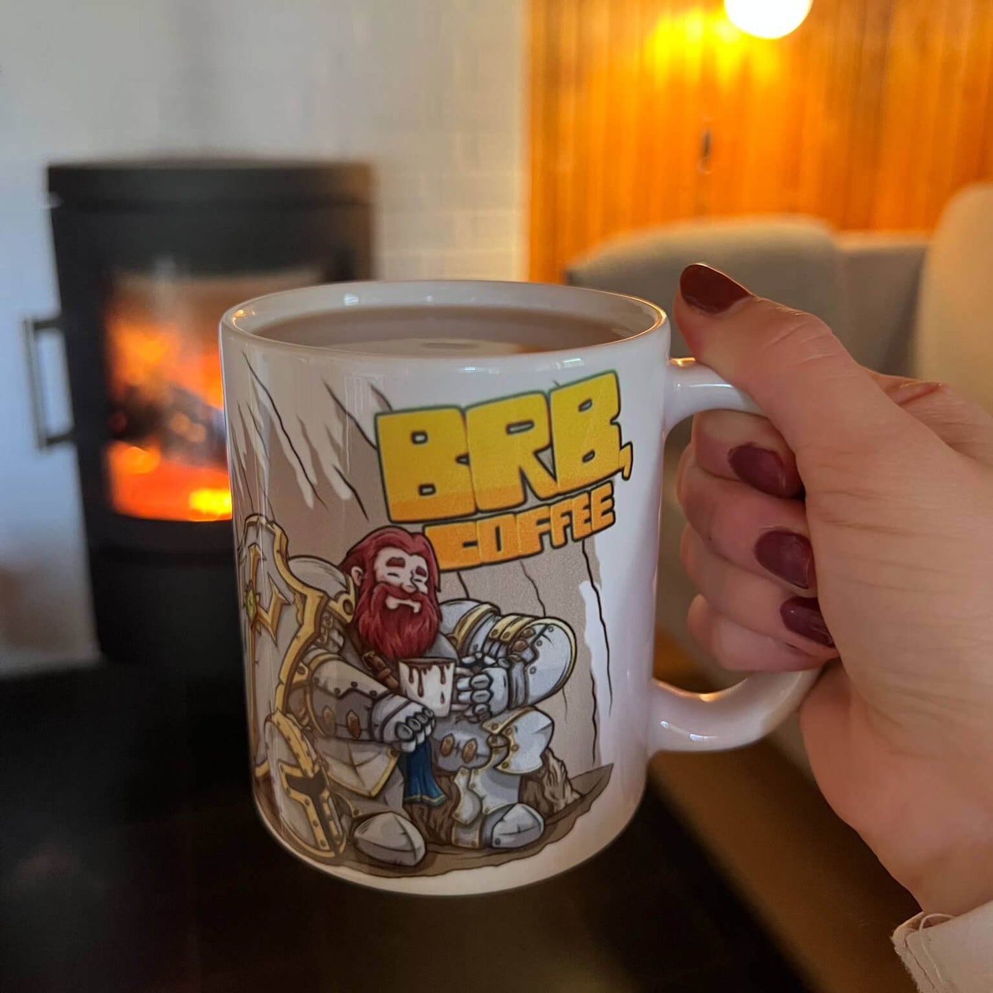 Person holding a mug with a cartoon knight and text 'BBB Coffee' design in front of a fireplace.