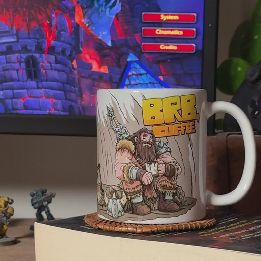 Video of the GeekyGearing "Barbarian’s Coffee Break" mug rotating on a coaster placed on top of a book, revealing the full wraparound design. A computer screen and Warhammer miniatures appear blurred in the background. Toward the end, coffee is poured into the mug from a moka pot.