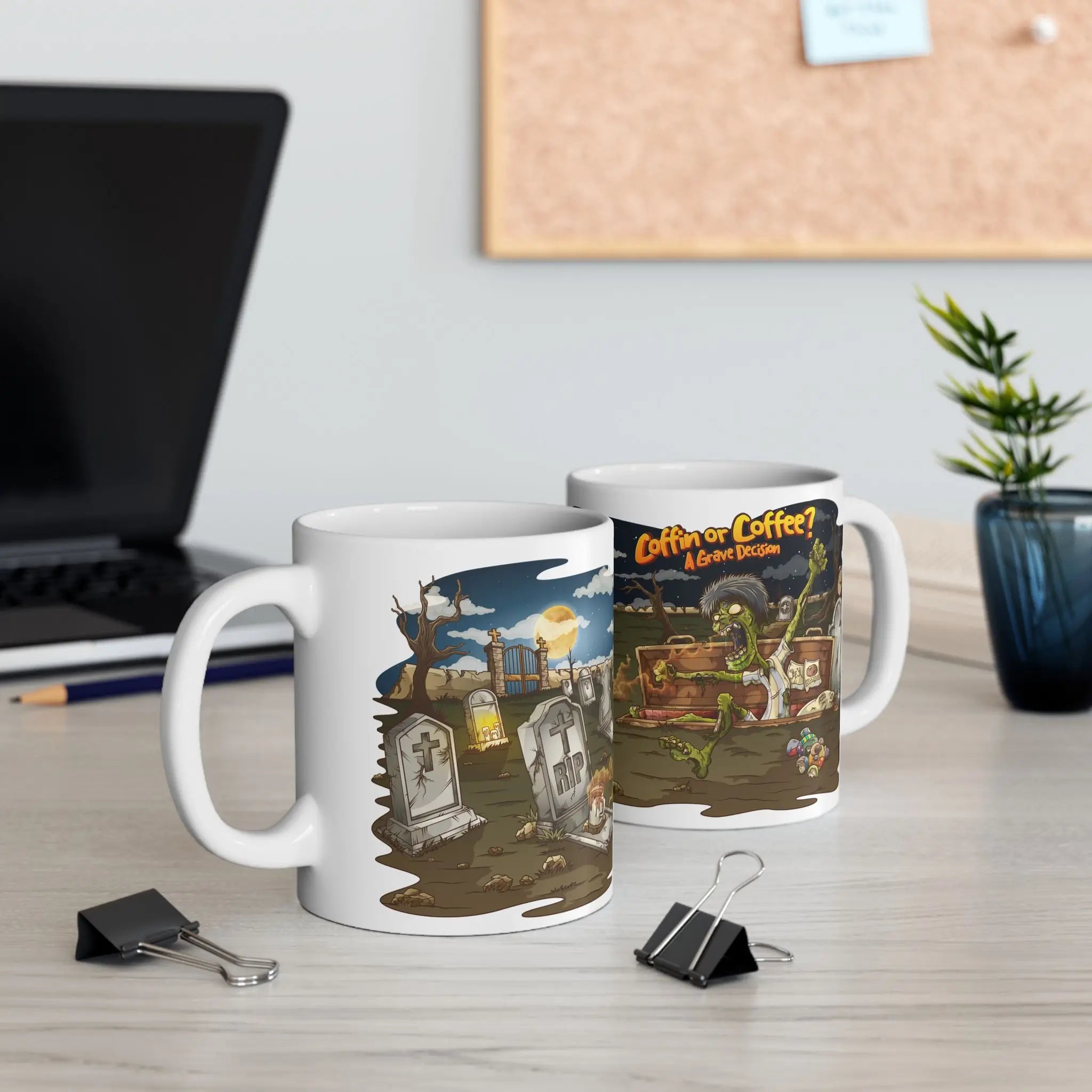 A halloween mug seen from both its sides featuring a zombie in a graveyard design. Mug is placed on a desk with a laptop and plant.