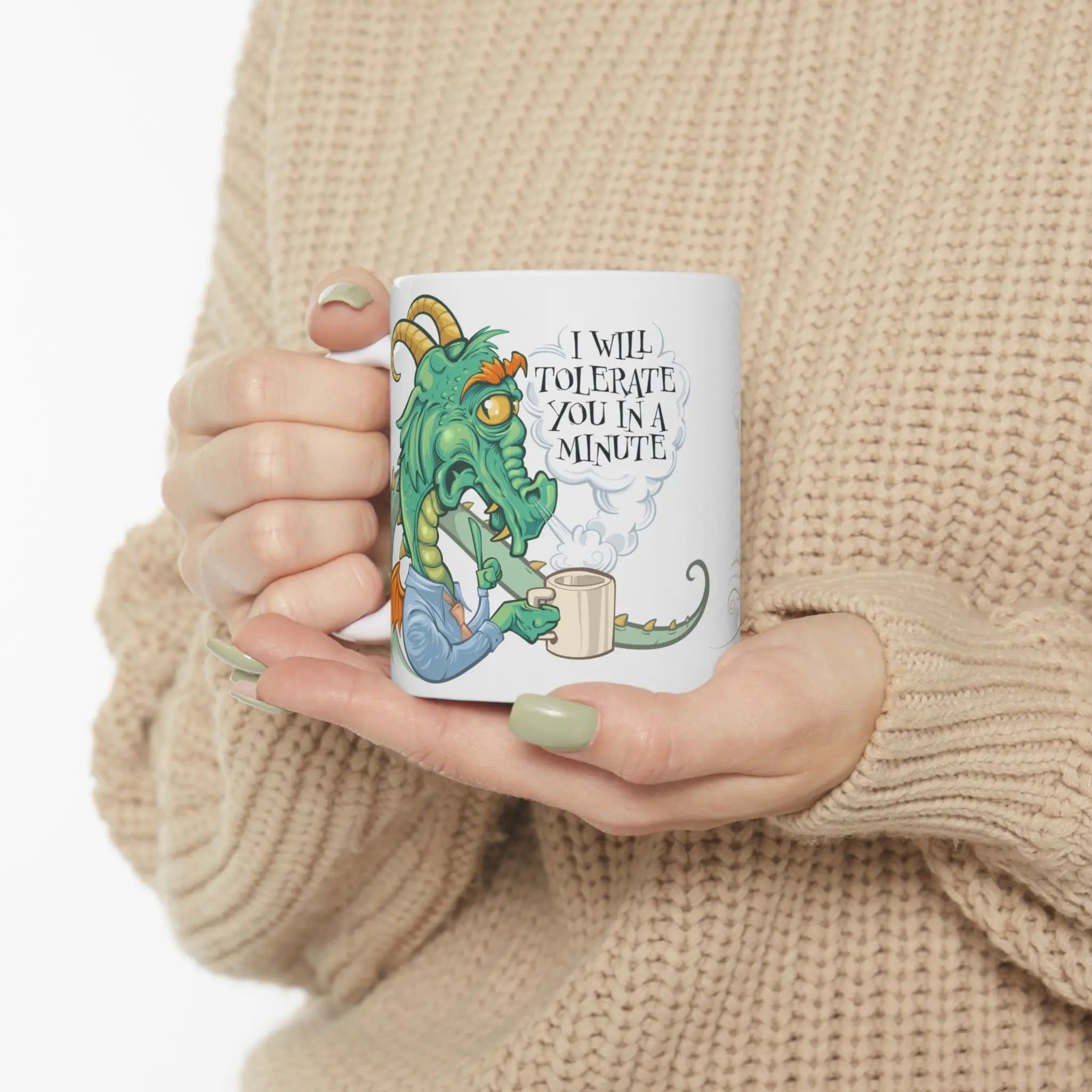 Person holding a mug with a dragon illustration and text, wearing a beige sweater.