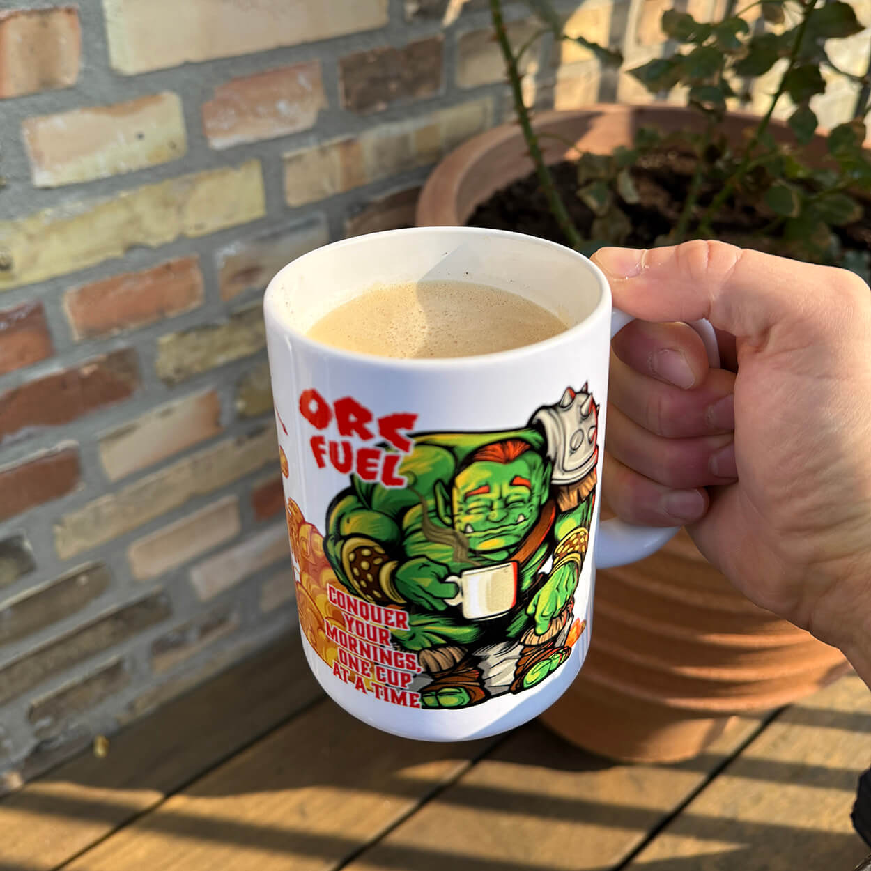 Hand holding a mug with a colorful design of a character and text, set against a brick wall and potted plant background.