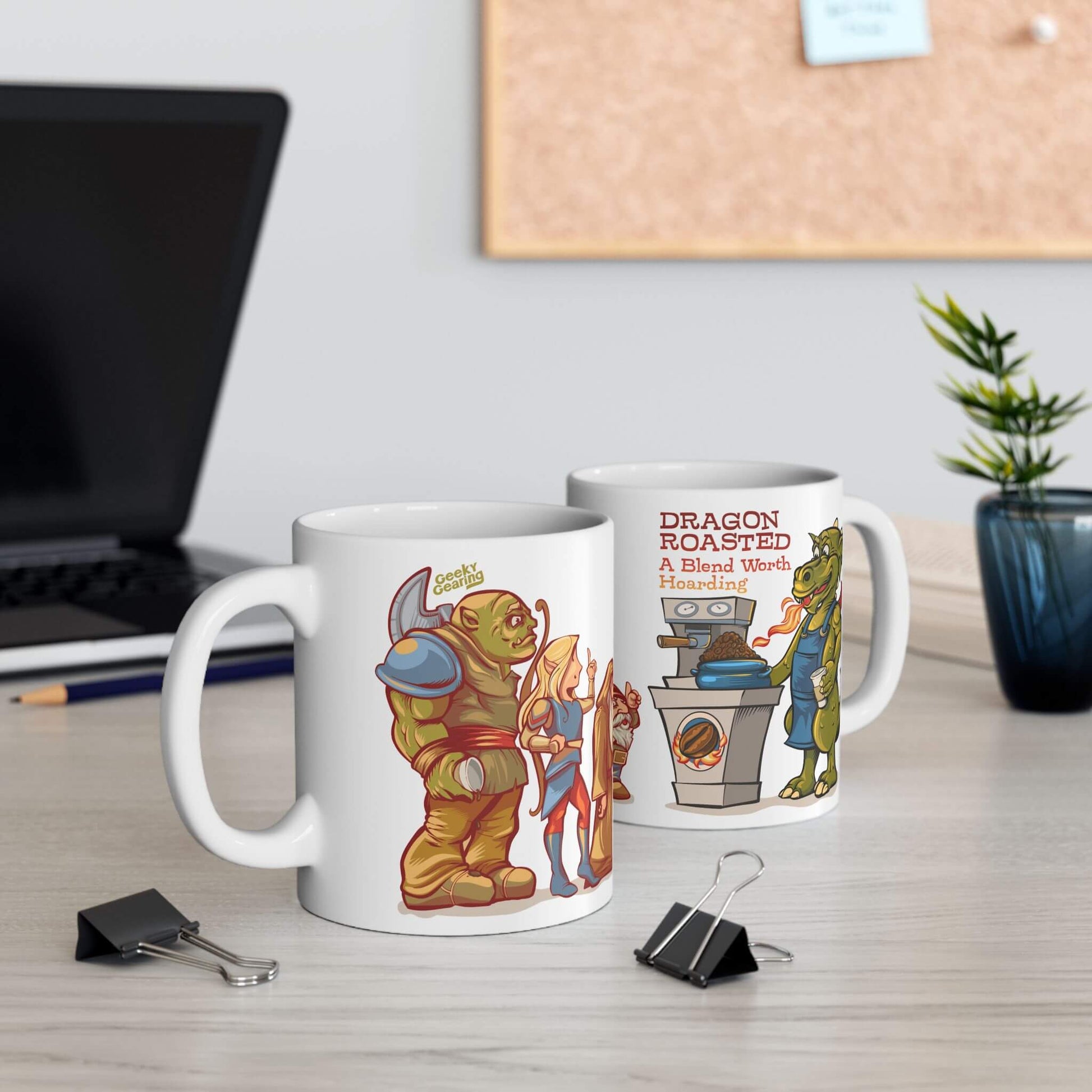 Two mugs with cartoon designs on a desk with a laptop and plant in the background.