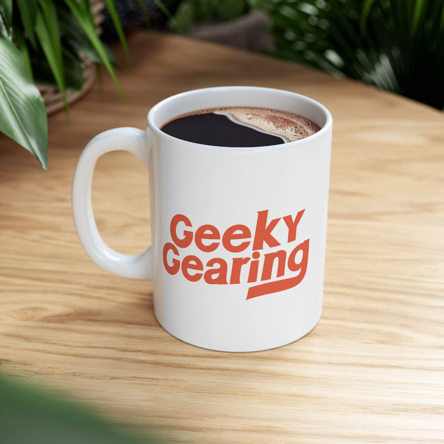 White mug with 'Geeky Gearing' logo on a wooden table with coffee.
