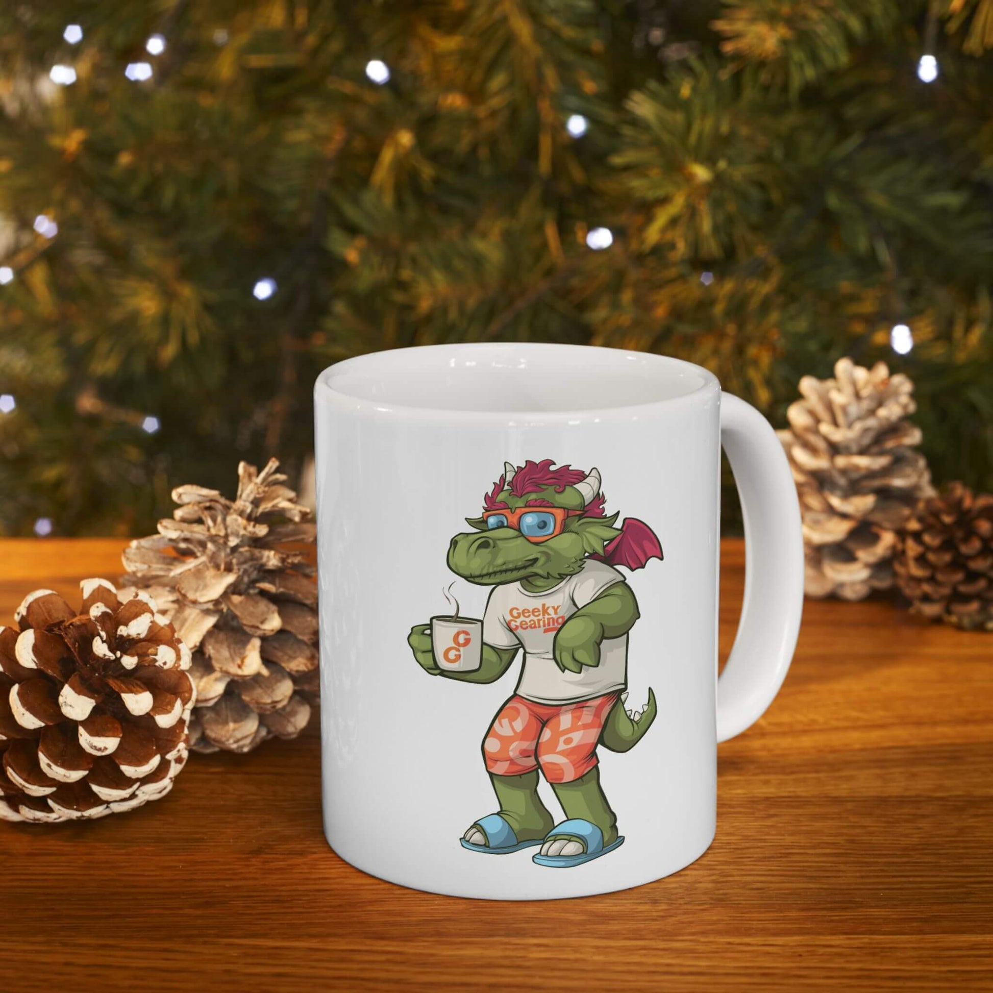 White mug with a cartoon dragon design holding a coffee cup, placed on a wooden surface with pinecones and a Christmas tree in the background.