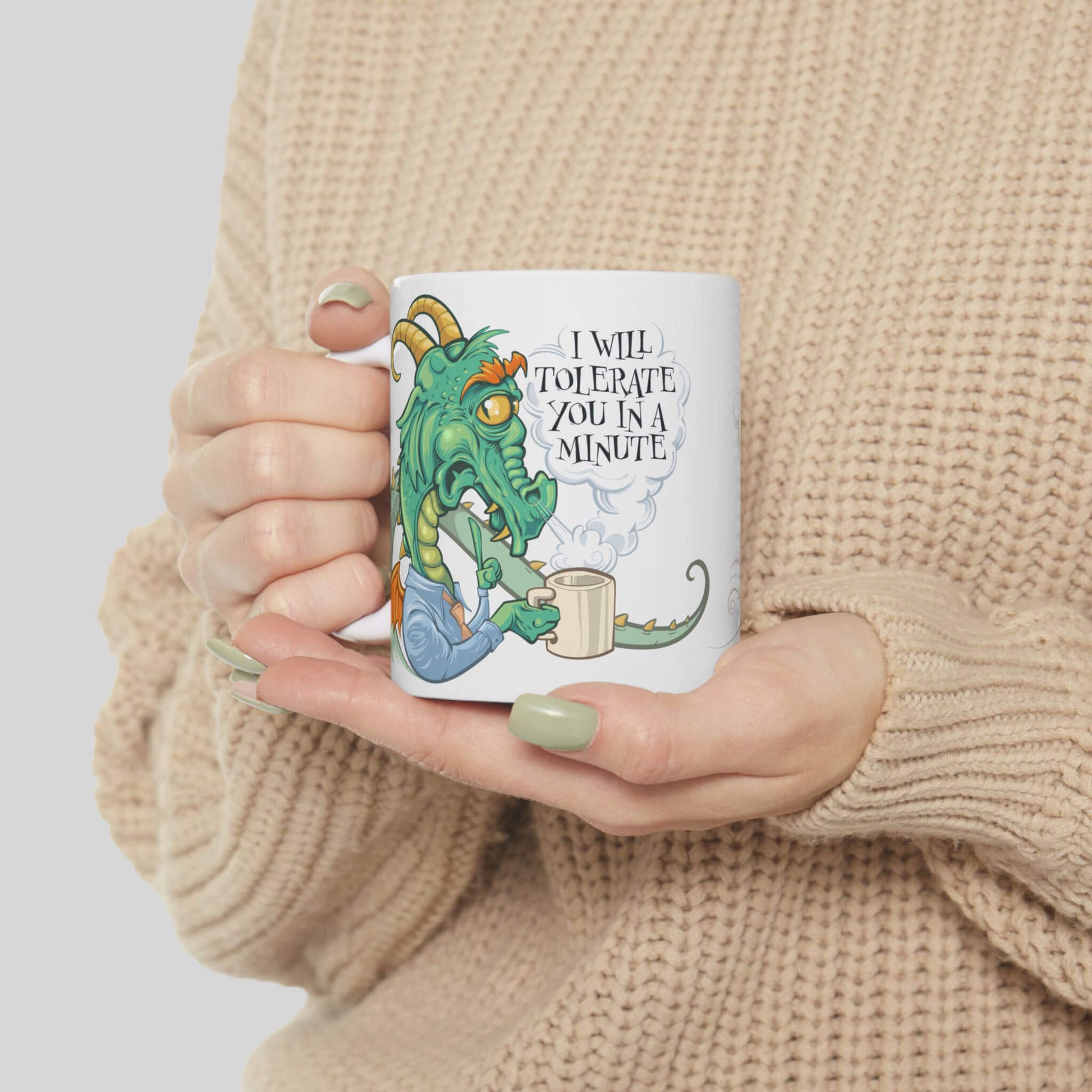 Person holding a mug with a dragon illustration and text, wearing a beige sweater.