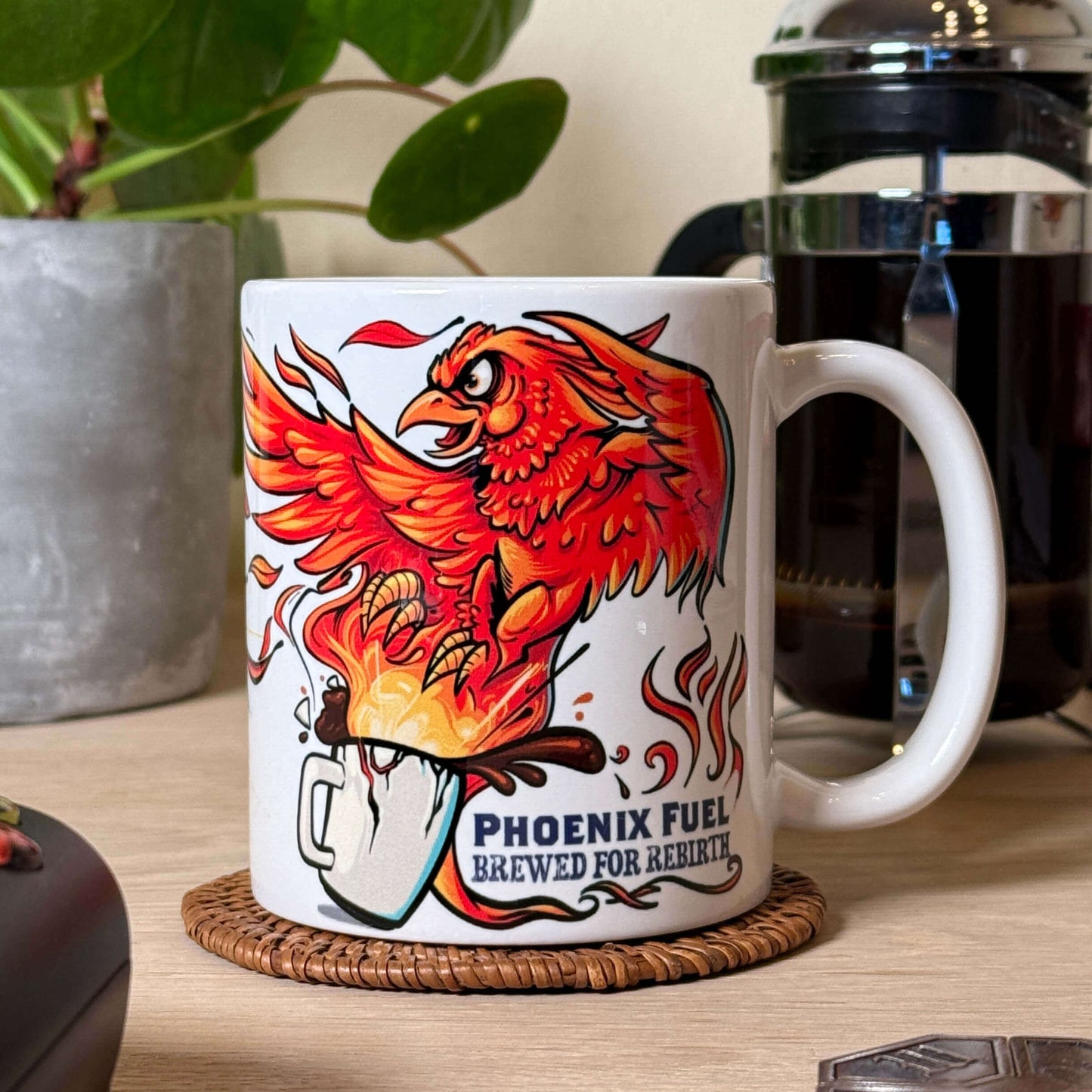 Mug with phoenix design and text 'Phoenix Fuel Brewed for Rebirth' on a table with a plant and coffee press in the background.