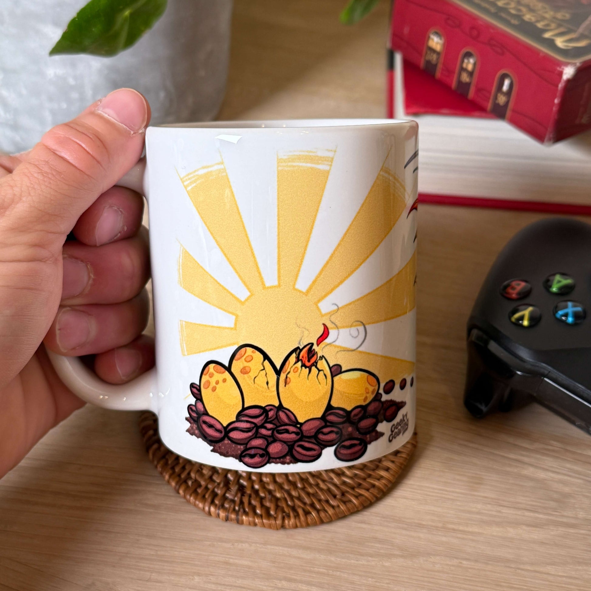Hand holds Phoenix Fuel mug showing phoenix hatching from egg in coffee beans with sunrise; game controller and plant in background.