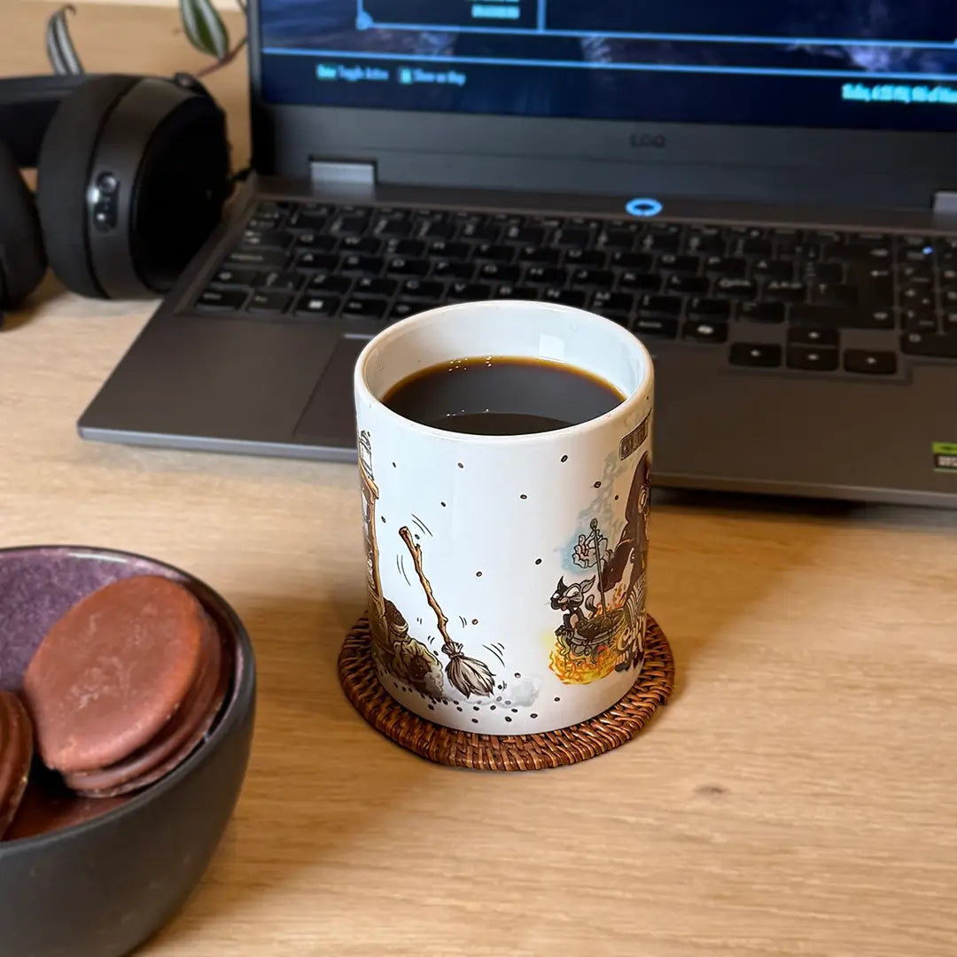 Front-angled view of an 11 oz white ceramic “Caffeine Is My Mana” fantasy mug filled with black coffee, resting on a woven coaster before a laptop and headphones on a wooden desk. The design wraps around to show a magical broom sweeping coffee grounds beside a bubbling wizard’s potion shelf. A bowl of macarons hints at a gamer’s breakfast. Perfect as a wizard mug, DnD gift, fantasy coffee cup or gamer gift.