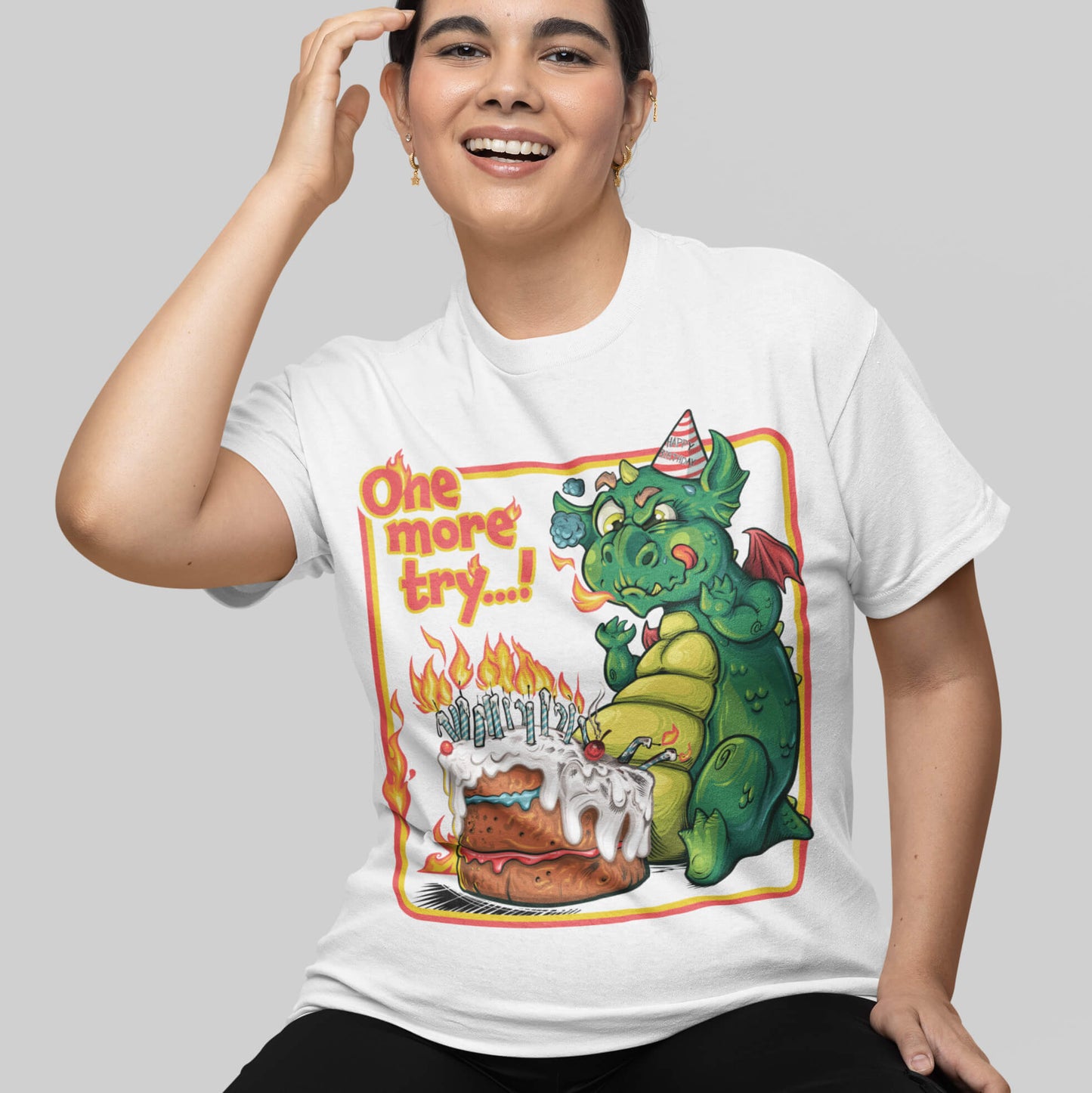 Person wearing a white t-shirt with a colorful graphic of a dragon and cake, text 'One more try...', on a plain background.