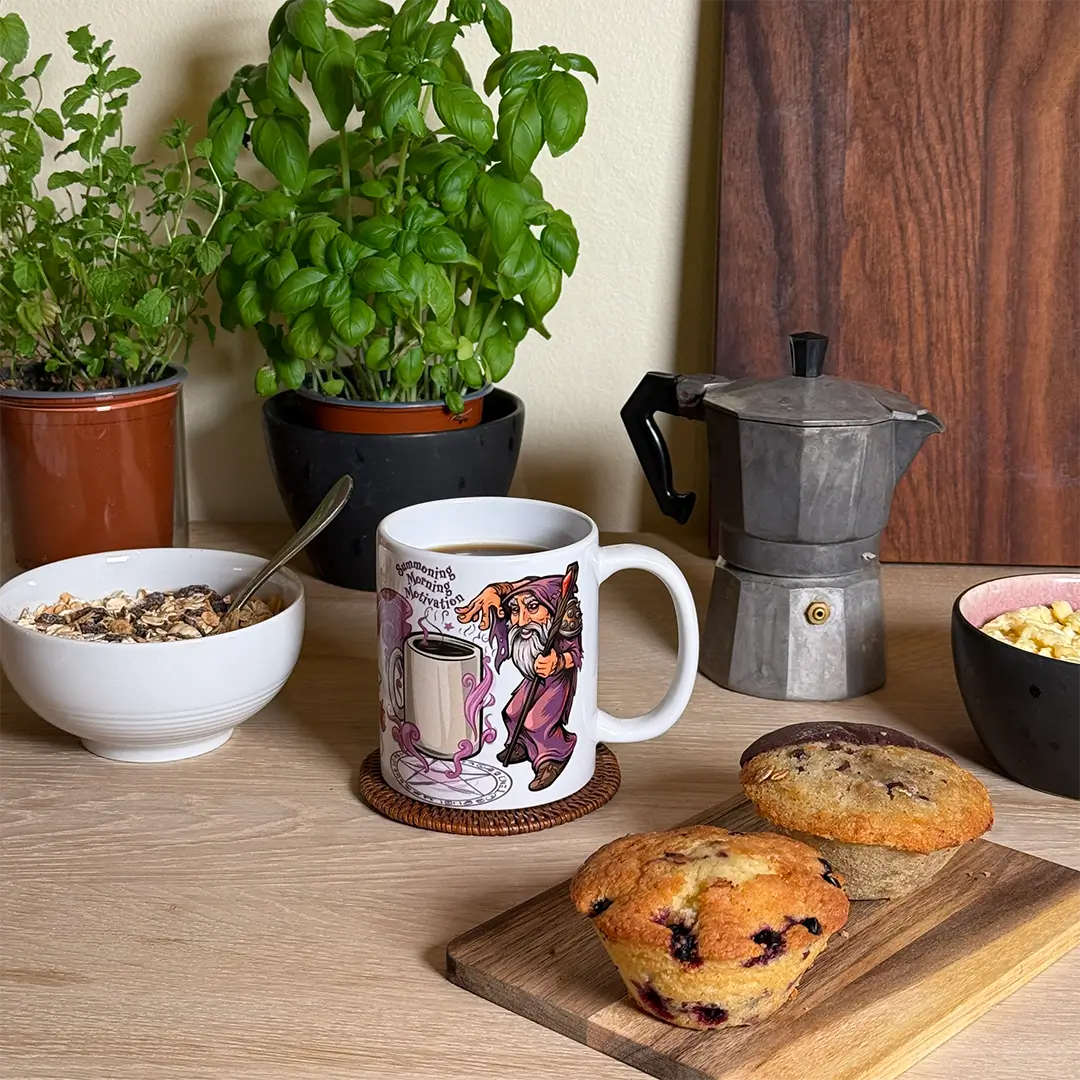 White 11 oz ceramic mug with warlock summoning coffee; moka pot and breakfast on table; text: “Summoning Morning Motivation”
